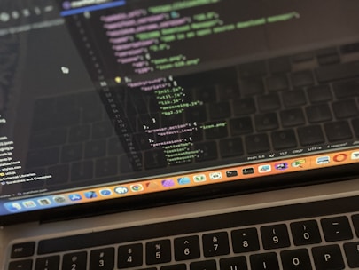 A close-up view of a laptop displaying code on the screen. The code editor is open on a dark-themed interface, showing lines of JavaScript with syntax highlighting. Below the screen, the keyboard of the laptop is partially visible. The laptop's dock at the bottom houses several application icons.