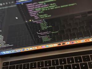 A close-up view of a laptop displaying code on the screen. The code editor is open on a dark-themed interface, showing lines of JavaScript with syntax highlighting. Below the screen, the keyboard of the laptop is partially visible. The laptop's dock at the bottom houses several application icons.