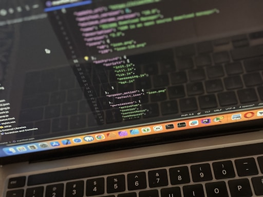 A close-up view of a laptop displaying code on the screen. The code editor is open on a dark-themed interface, showing lines of JavaScript with syntax highlighting. Below the screen, the keyboard of the laptop is partially visible. The laptop's dock at the bottom houses several application icons.