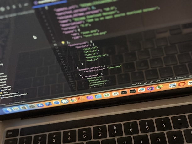 A close-up view of a laptop displaying code on the screen. The code editor is open on a dark-themed interface, showing lines of JavaScript with syntax highlighting. Below the screen, the keyboard of the laptop is partially visible. The laptop's dock at the bottom houses several application icons.
