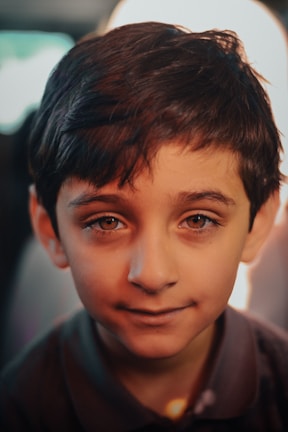 A young boy with dark hair and soulful eyes, gazing directly at the camera with a gentle smile. The lighting creates warm tones on his face, and there is a soft, blurred background.