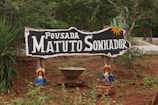 A rustic wooden sign with the text 'Pousada Matuto Sonhador' in white letters is prominently displayed in a natural, outdoor setting. The sign features decorative elements, including small figures of people in traditional attire positioned on either side at the base. The background consists of trees and lush greenery, suggesting a rural or forested location.