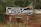 A rustic wooden sign with the text 'Pousada Matuto Sonhador' in white letters is prominently displayed in a natural, outdoor setting. The sign features decorative elements, including small figures of people in traditional attire positioned on either side at the base. The background consists of trees and lush greenery, suggesting a rural or forested location.