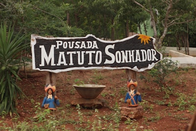 A rustic wooden sign with the text 'Pousada Matuto Sonhador' in white letters is prominently displayed in a natural, outdoor setting. The sign features decorative elements, including small figures of people in traditional attire positioned on either side at the base. The background consists of trees and lush greenery, suggesting a rural or forested location.