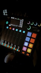 A digital audio mixing console with colorful buttons and sliders. The console has a small display screen showing various controls and settings. The ambient lighting highlights the vibrant colors of the buttons.