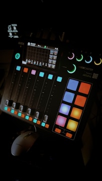 A digital audio mixing console with colorful buttons and sliders. The console has a small display screen showing various controls and settings. The ambient lighting highlights the vibrant colors of the buttons.
