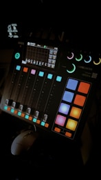 A digital audio mixing console with colorful buttons and sliders. The console has a small display screen showing various controls and settings. The ambient lighting highlights the vibrant colors of the buttons.