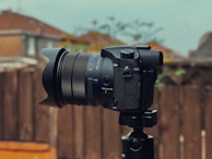 A professional camera is mounted on a tripod outdoors, with a blurred background of wooden fencing and rooftops. The camera features a large lens with detailed branding visible and several dials and buttons.
