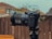 A professional camera is mounted on a tripod outdoors, with a blurred background of wooden fencing and rooftops. The camera features a large lens with detailed branding visible and several dials and buttons.