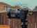 A professional camera is mounted on a tripod outdoors, with a blurred background of wooden fencing and rooftops. The camera features a large lens with detailed branding visible and several dials and buttons.