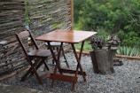 A wooden table and chair set placed on a gravel surface, surrounded by a rustic wooden fence made from tree branches. The background features lush green foliage and small plants near the fence. The setting gives a natural and serene atmosphere.