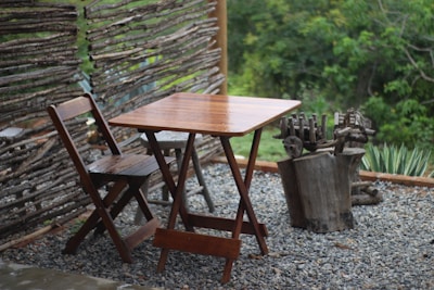 A wooden table and chair set placed on a gravel surface, surrounded by a rustic wooden fence made from tree branches. The background features lush green foliage and small plants near the fence. The setting gives a natural and serene atmosphere.