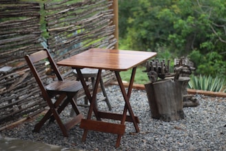 A wooden table and chair set placed on a gravel surface, surrounded by a rustic wooden fence made from tree branches. The background features lush green foliage and small plants near the fence. The setting gives a natural and serene atmosphere.
