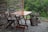 A wooden table and chair set on a small gravel patio beside a rustic fence made from thin tree branches. A tree stump sculpture sits nearby, with lush greenery in the background.
