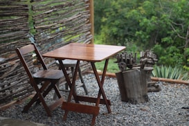 A wooden table and chair set on a small gravel patio beside a rustic fence made from thin tree branches. A tree stump sculpture sits nearby, with lush greenery in the background.