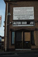 A corner of a building with the name 'Alter Ego Barber Shop' displayed. Above the shop name, several signs are visible, one addressing break-ins and another offering an appointment system. The facade shows the shop's opening times, and parts of an adjacent street sign and buildings are also in view.