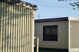Two prefabricated buildings with corrugated metal siding stand side by side. The buildings have flat roofs and small windows. Shadows from nearby trees create patterns on the walls, and power lines are visible in the background. The scene is illuminated by natural daylight.