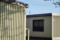 Two prefabricated buildings with corrugated metal siding stand side by side. The buildings have flat roofs and small windows. Shadows from nearby trees create patterns on the walls, and power lines are visible in the background. The scene is illuminated by natural daylight.