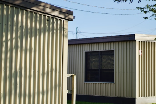Two prefabricated buildings with corrugated metal siding stand side by side. The buildings have flat roofs and small windows. Shadows from nearby trees create patterns on the walls, and power lines are visible in the background. The scene is illuminated by natural daylight.