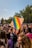 A vibrant crowd gathers at an outdoor event, with numerous individuals in colorful attire and some holding rainbow flags. A prominently displayed sign reads, 'I'M NOT AS GAY AS I LOOK (I'M MUCH GAYER)'. The setting is bright with clear skies, suggesting a festive and inclusive atmosphere.