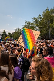 A vibrant crowd gathers at an outdoor event, with numerous individuals in colorful attire and some holding rainbow flags. A prominently displayed sign reads, 'I'M NOT AS GAY AS I LOOK (I'M MUCH GAYER)'. The setting is bright with clear skies, suggesting a festive and inclusive atmosphere.