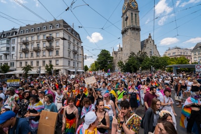 A large crowd of people, many wearing rainbow-colored attire and holding banners, are gathered in a city square with a tall clock tower in the background. Overhead wires for trams are visible, and the buildings around have European architectural styles. The mood appears vibrant and celebratory.