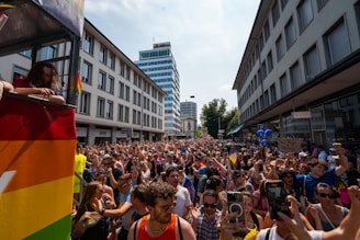 A large crowd gathered in an urban street surrounded by tall buildings, participating in a parade with vibrant rainbow flags and balloons. The atmosphere is festive with people wearing colorful clothing, holding up phones, and cheering. A sign offering free hugs and kisses is visible.