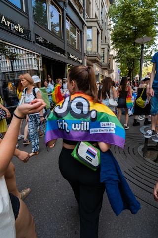 People are walking on a street lined with shops, including Van Cleef & Arpels. The scene includes a person draped in a rainbow flag with a message in German. Others around are also holding pride-related items, and the group appears to be participating in a pride march or event. The setting is urban with trees and buildings lining the street.