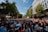 A large crowd of people gathers on a city street, participating in an event under a clear blue sky. Many attendees are carrying rainbow flags and wearing colorful clothing, indicating a pride parade or celebration. Surrounding the crowd are tall buildings and trees lining the street.