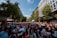 A large crowd of people gathers on a city street, participating in an event under a clear blue sky. Many attendees are carrying rainbow flags and wearing colorful clothing, indicating a pride parade or celebration. Surrounding the crowd are tall buildings and trees lining the street.