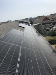 A row of solar panels is installed on a sloped rooftop, extending into the distance. The background features a suburban neighborhood with multiple houses of various colors and sizes. The sky appears clear with subtle cloud formations.