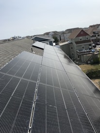 A row of solar panels is installed on a sloped rooftop, extending into the distance. The background features a suburban neighborhood with multiple houses of various colors and sizes. The sky appears clear with subtle cloud formations.