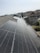 A row of solar panels is installed on a sloped rooftop, extending into the distance. The background features a suburban neighborhood with multiple houses of various colors and sizes. The sky appears clear with subtle cloud formations.