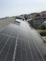 A row of solar panels is installed on a sloped rooftop, extending into the distance. The background features a suburban neighborhood with multiple houses of various colors and sizes. The sky appears clear with subtle cloud formations.
