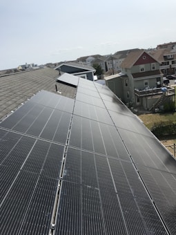 A row of solar panels is installed on a sloped rooftop, extending into the distance. The background features a suburban neighborhood with multiple houses of various colors and sizes. The sky appears clear with subtle cloud formations.