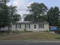 A single-story house with beige siding and dark green shutters. Solar panels are installed on the roof. There's a grassy front yard with some plants and small shrubs. A utility vehicle with 'Sunrun' branding is parked to the right, along with another car.