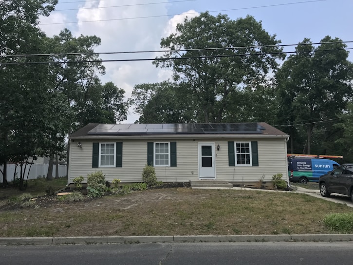 A single-story house with beige siding and dark green shutters. Solar panels are installed on the roof. There's a grassy front yard with some plants and small shrubs. A utility vehicle with 'Sunrun' branding is parked to the right, along with another car.