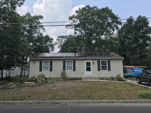 A single-story house with beige siding and dark green shutters. Solar panels are installed on the roof. There's a grassy front yard with some plants and small shrubs. A utility vehicle with 'Sunrun' branding is parked to the right, along with another car.