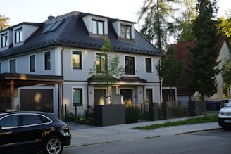 A modern, two-story house with a dark roof and light-colored walls is situated on a quiet residential street. In front of the house, there is a driveway with a black car parked, and some greenery and small trees surround the property.