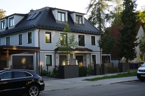 A modern, two-story house with a dark roof and light-colored walls is situated on a quiet residential street. In front of the house, there is a driveway with a black car parked, and some greenery and small trees surround the property.