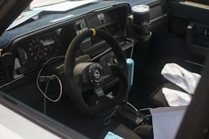 The interior of a car featuring a steering wheel with a racing emblem, dashboard, and gauges. There are items scattered inside, including a drink holder with a cup, a cable hanging over the steering column, and cloth materials on the passenger seat.