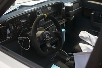 The interior of a car featuring a steering wheel with a racing emblem, dashboard, and gauges. There are items scattered inside, including a drink holder with a cup, a cable hanging over the steering column, and cloth materials on the passenger seat.