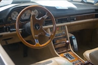 A classic car interior featuring a wooden steering wheel and dashboard accents. The dashboard includes an array of buttons and dials, with retro-styled instruments. A vintage mobile phone is mounted next to the gear shift. The seats and interior are finished in beige leather.