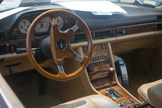 A classic car interior featuring a wooden steering wheel and dashboard accents. The dashboard includes an array of buttons and dials, with retro-styled instruments. A vintage mobile phone is mounted next to the gear shift. The seats and interior are finished in beige leather.