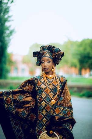 A person wearing an elaborate and colorful traditional outfit adorned with intricate geometric patterns. The attire includes a headdress and layers of beaded jewelry in vibrant hues. The background is blurred, featuring greenery and an indistinct structure.