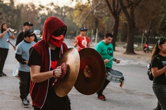 A group of people appears to be performing in an outdoor setting, possibly a park. One individual in the foreground is playing cymbals, wearing a red hoodie and a red face mask. Others are playing drums, brass instruments, and are dressed casually. The scene suggests a lively and energetic atmosphere.