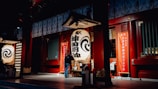 A warmly lit traditional Japanese lantern hangs prominently in front of an ornate building with red pillars and decorative elements. The setting suggests a sense of cultural heritage and celebration, with red banners inscribed with Japanese characters adding to the ambiance.