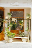 A storefront decorated with green plants in various pots and hanging containers. The window is framed with wooden accents and features a person inside, arranging flowers. The facade includes a decorative snail motif.