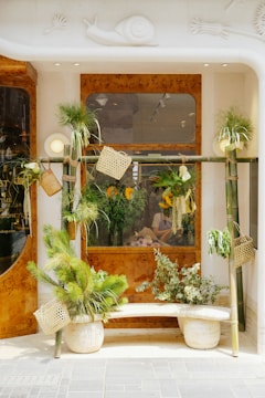 A storefront decorated with green plants in various pots and hanging containers. The window is framed with wooden accents and features a person inside, arranging flowers. The facade includes a decorative snail motif.