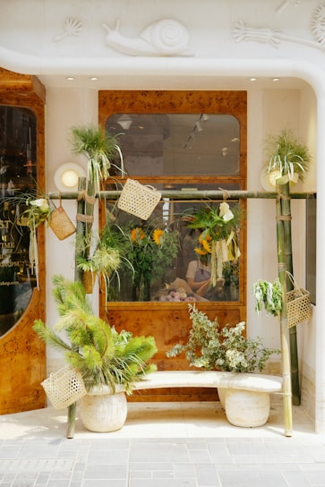 A storefront decorated with green plants in various pots and hanging containers. The window is framed with wooden accents and features a person inside, arranging flowers. The facade includes a decorative snail motif.